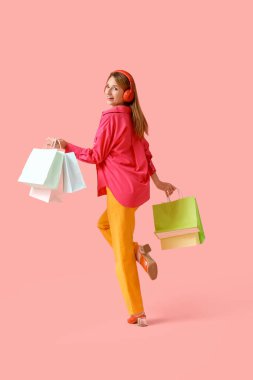 Pembe arka planda alışveriş torbaları olan kulaklıklı güzel, genç ve mutlu bir kadın.