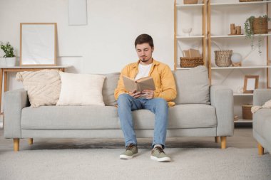 Genç adam evdeki kanepede kitap okuyor.