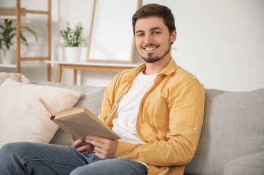 Genç adam evdeki kanepede kitap okuyor.