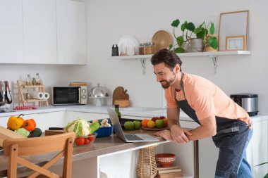 Mutfakta dizüstü bilgisayarı olan yakışıklı sakallı adam.