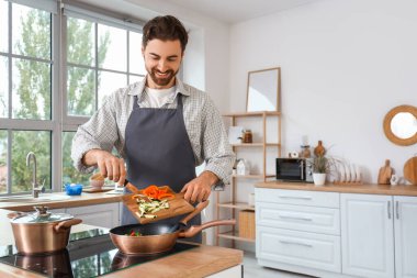 Yakışıklı sakallı adam mutfakta sebzeleri kızartma tavasına koyuyor.
