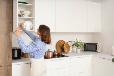 Genç bir kadın mutfaktaki açık dolaptan cam alıyor.