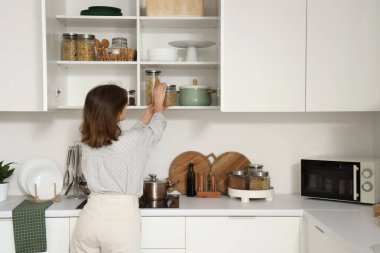 Genç bir kadın mutfaktaki raftan kavanozu alıyor.