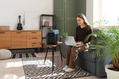 Genç bir kadın evdeki koltukta taşınabilir elektrik santraliyle dizüstü bilgisayarı şarj ediyor.