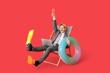 Şişirilebilir yüzüklü ve yüzgeçli bir ofis çalışanı kırmızı arka planda dizüstü bilgisayarıyla güverte koltuğunda oturuyor.