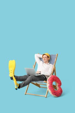 Ofis çalışanı dizüstü bilgisayarla güvertede oturuyor ve mavi arka planda tatilin hayalini kuruyor.