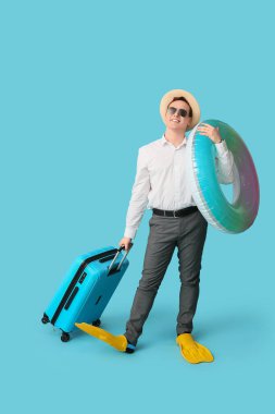 Şişme yüzüklü, bavullu ve paletli bir ofis çalışanı mavi arka planda tatil hayali kuruyor.