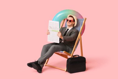 Ofis çalışanı elinde şişirilebilir yüzük ve gazeteyle güverte koltuğunda oturmuş pembe arka planda tatil hayali kuruyor.