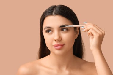 Young woman correcting shape of her eyebrows on brown background clipart