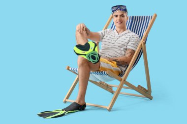 Mavi arka planda şnorkelli ve paletli genç adam güverte koltuğunda oturuyor.