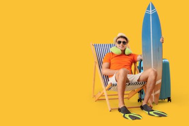 Sörf tahtası olan genç adam sarı arka planda iskemlede oturuyor.