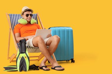 Dizüstü bilgisayarlı genç adam güvertede oturuyor ve sarı arka planda plaj aksesuarları var.