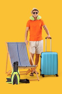 Young man with suitcase and deck chair on yellow background