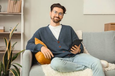 E-okuyuculu genç adam evdeki kanepede oturuyor.