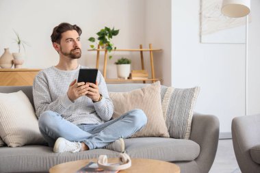 E-okuyuculu genç adam evdeki kanepede oturuyor.