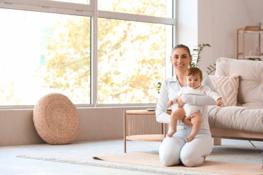 Küçük bebeğiyle evde yoga minderinde oturan sportif genç bir anne.