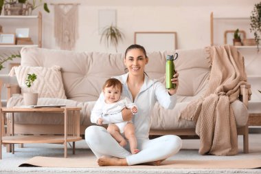 Küçük bebeği ve su şişesiyle evde yoga minderinde oturan sportif genç anne.