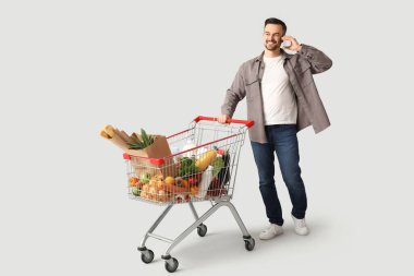 Alışveriş arabalı mutlu genç adam gri arka planda cep telefonuyla konuşuyor.