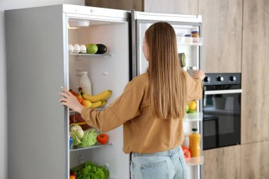 Mutfakta açık buzdolabının yanında genç bir kadın var.