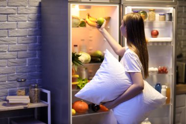 Yastıklı genç bir kadın geceleri mutfaktaki buzdolabından elma alıyor.