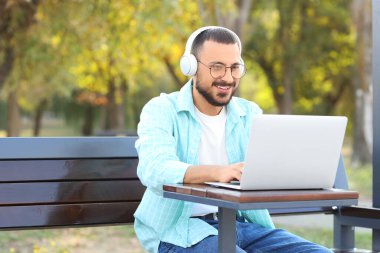Kulaklıklı genç adam parkta bankta dizüstü bilgisayar kullanıyor.