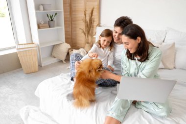 Pomeranya köpeği ve dizüstü bilgisayarı olan mutlu bir aile yatak odasında oturuyor.