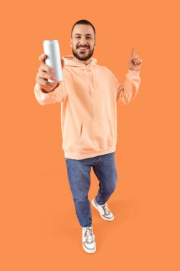 Elinde soda olan genç adam turuncu arka plandaki bir şeyi işaret ediyor.