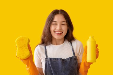 Sarı arka planda sünger ve deterjanı olan genç bir kadın.