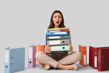 Shocked young businesswoman with folders sitting on grey background clipart