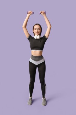 Kulaklıklı ve dambılı genç bayan antrenör leylak arka planı üzerinde egzersiz yapıyor.