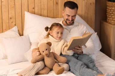 Mutlu baba ve küçük kızı oyuncak ayıyla evde kış akşamı yatakta kitap okuyorlar.