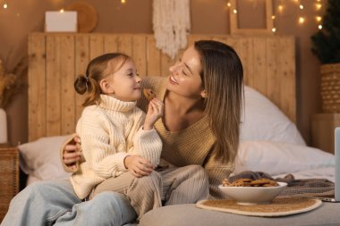 Mutlu anne ve küçük kızı kışlık giysiler içinde evde kış akşamı yatakta kurabiye yiyorlar.