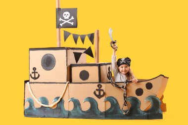 Korsan gibi giyinmiş şirin küçük kız sarı arka planda karton gemide kılıçla oynuyor.