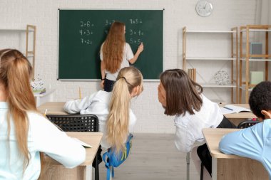 Sınıftaki küçük kızlar ders sırasında öğretmenlerinin arkasından fısıldaşıyorlar.