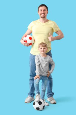 Sevimli küçük çocuk ve futbol toplarıyla genç babası mavi arka planda başparmak kaldırıyor.