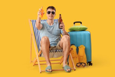 Bira ve güneş kremi sürmüş genç adam sarı arka planda iskemlede oturuyor.