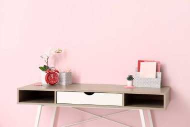 Alarmlı saati, ev bitkisi ve rahat ofisteki pembe duvarın yanındaki defterleri olan şık bir iş yeri.