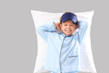 Pijamalı, tatlı, mutlu bir çocuk. Gri arka planda uyku maskesi ve yastığı var.