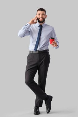 Gri arka planda cep telefonuyla konuşan bir fincan kahveyle yakışıklı, genç ve mutlu bir iş adamı.