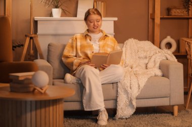 Evdeki kanepede kitap okuyan güzel, genç ve mutlu bir kadın.