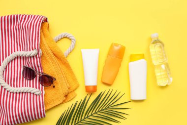 Sarı zemin üzerinde güneş kremi, palmiye yaprağı, çanta ve güneş gözlüğü olan kompozisyon