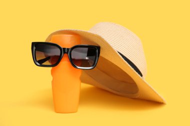 Sarı arka planda güneş kremi, şapka ve güneş gözlüğü olan kompozisyon