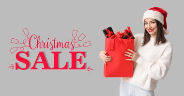 Noel Baba şapkalı güzel bir kadın gri arka planda hediye kutuları olan bir alışveriş çantası tutuyor. Noel indirimi