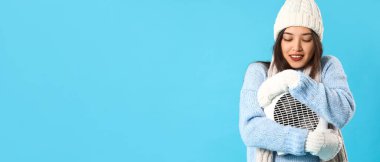 Kışın giyinmiş genç Asyalı kadın açık mavi arka planda elektrikli fan ısıtıcısına sarılıyor ve mesaj için yeri var.