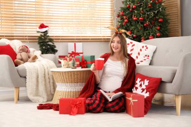 Geyik boynuzları, hediye kutuları ve Noel için dekore edilmiş odada kakao okuyan genç hamile kadın.