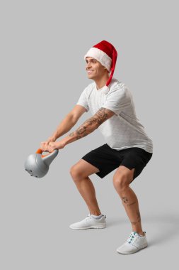 Noel Baba şapkalı genç adam gri arka planda kettlebell eğitimi alıyor.