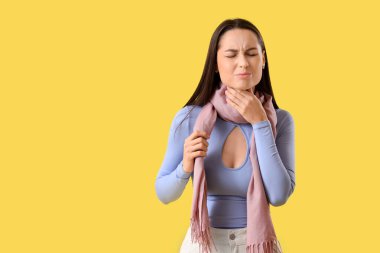 Sarı arka planda boğazı ağrıyan fularlı genç kadın.