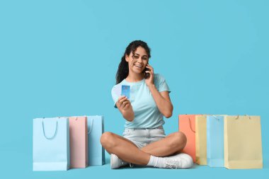 Mavi arka planda cep telefonu, kredi kartı ve alışveriş torbalarıyla oturan genç bir kadın.