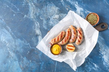 Lezzetli ızgara sosis, soğan ve mavi arka planda bir kase hardal ile pişirme kağıdı.