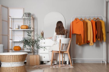 Güz kıyafetleriyle soyunma odasının penceresinin yanında makyaj yapan genç bir kadın.